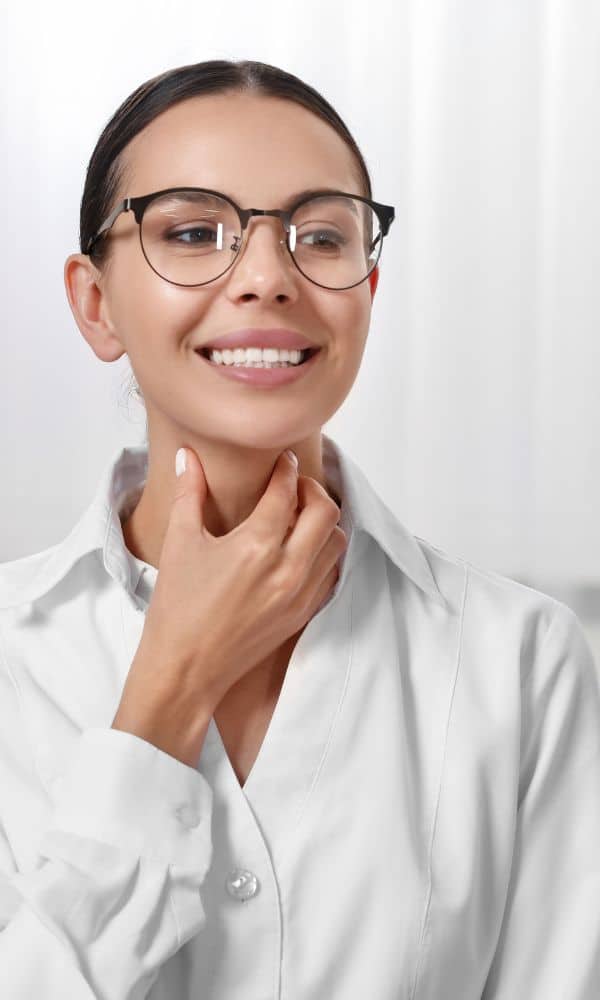 Portrait d’une femme en blouse blanche touchant sa gorge en souriant, illustrant un exercice de phonation ou une démonstration de technique vocale, souvent utilisé en orthophonie.
