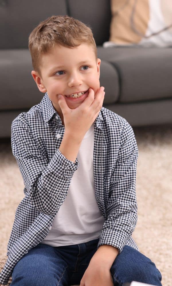 Un jeune garçon souriant assis au sol, la main posée sur sa joue, reproduit un mouvement de bouche dans un contexte ludique, illustrant une séance de langage ou d’orthophonie.