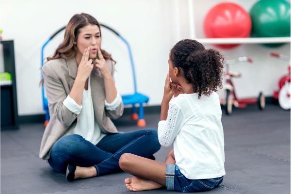 Une orthophoniste assise en tailleur face à une enfant imite un mouvement de bouche, illustrant une séance de rééducation du langage dans un cadre bienveillant et adapté aux enfants.