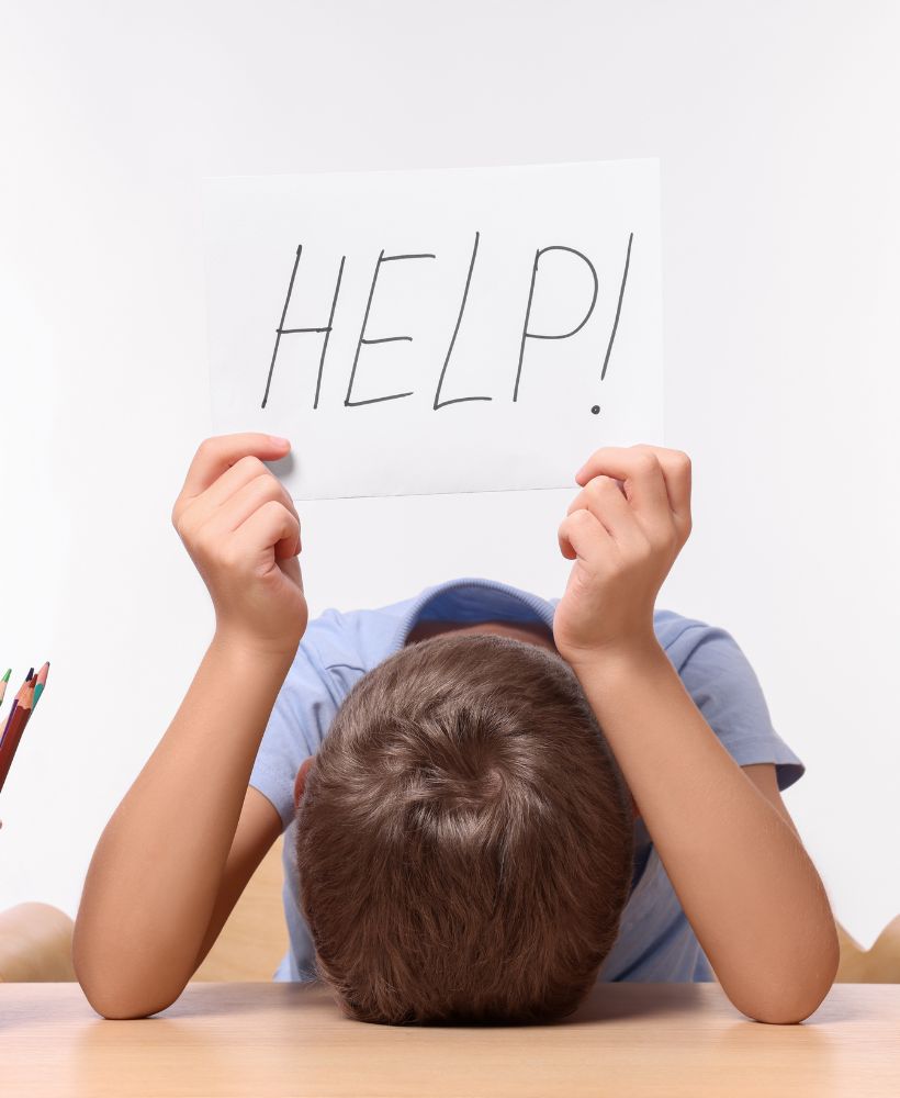Un enfant assis à un bureau, la tête posée sur les bras, tenant une feuille avec le mot 